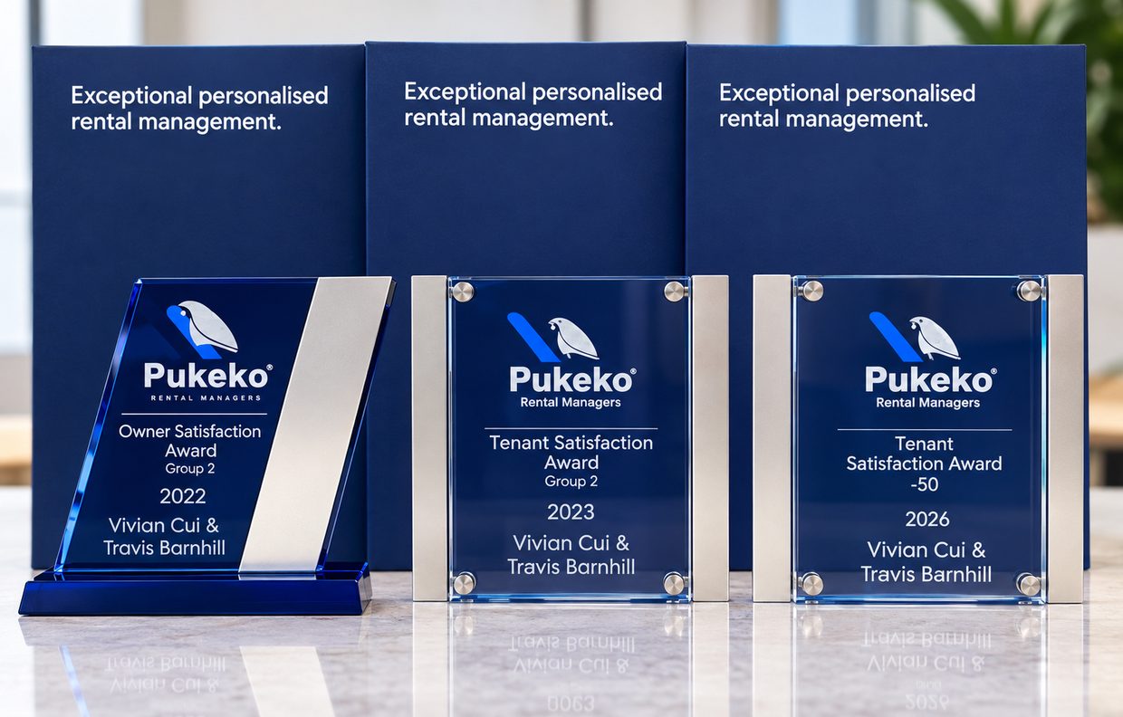 Three blue glass Pukeko awards for Vivian Cui and Travis Barnhill, Owner Satisfaction Award 2022, Tenant Satisfaction Award 2023 and Tenant Satisfaction Award 2026, in front of navy folders reading Exceptional personalised rental management.