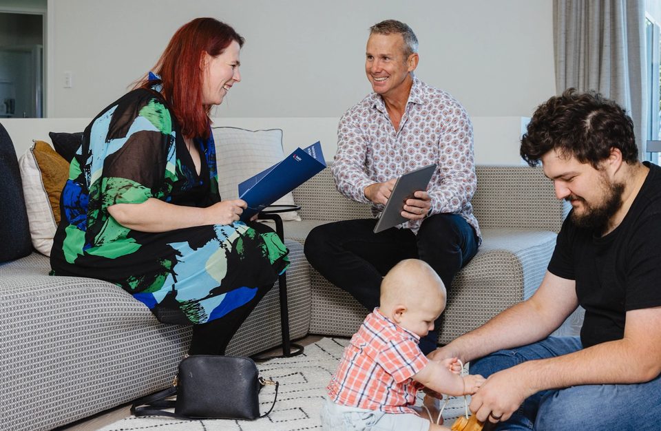A Pukeko rental manager with a tablet talking to a young couple and their baby in a lounge, the woman holding a blue Pukeko folder.