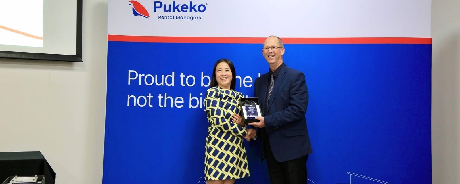 Vivian Cui receiving a Pukeko Rental Managers award on stage in front of a blue Pukeko banner that reads Proud to be the best, not the biggest.