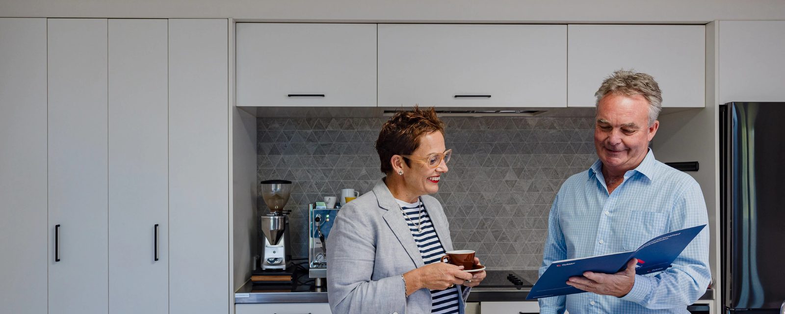 A Pukeko rental manager with a coffee cup laughing in a white kitchen beside an espresso machine.