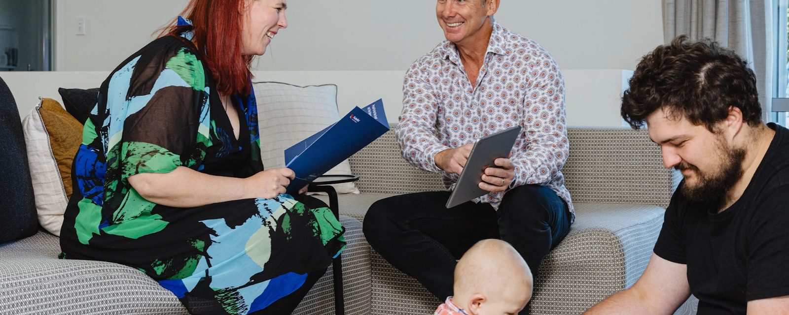 A Pukeko rental manager with a tablet talking to a young couple and their baby in a lounge, the woman holding a blue Pukeko folder.