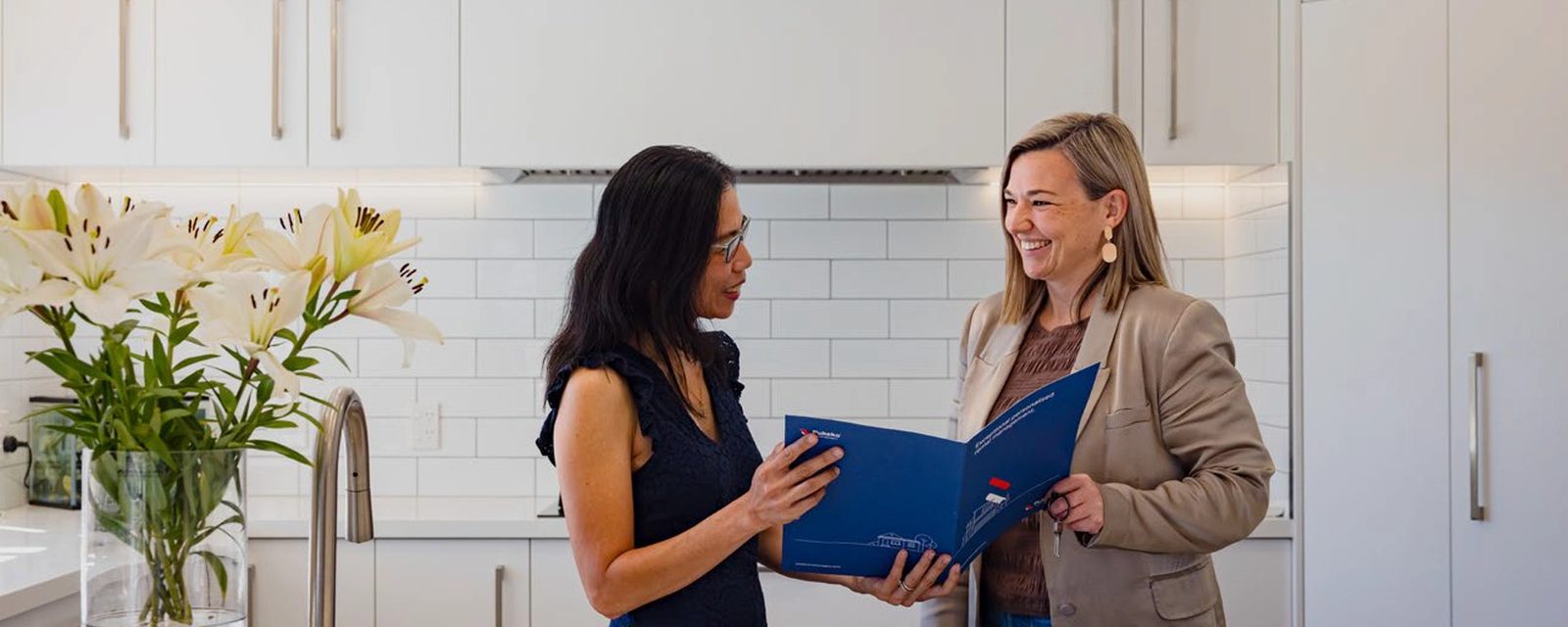 A property owner and a Pukeko rental manager reading through a blue management folder together in a white kitchen.