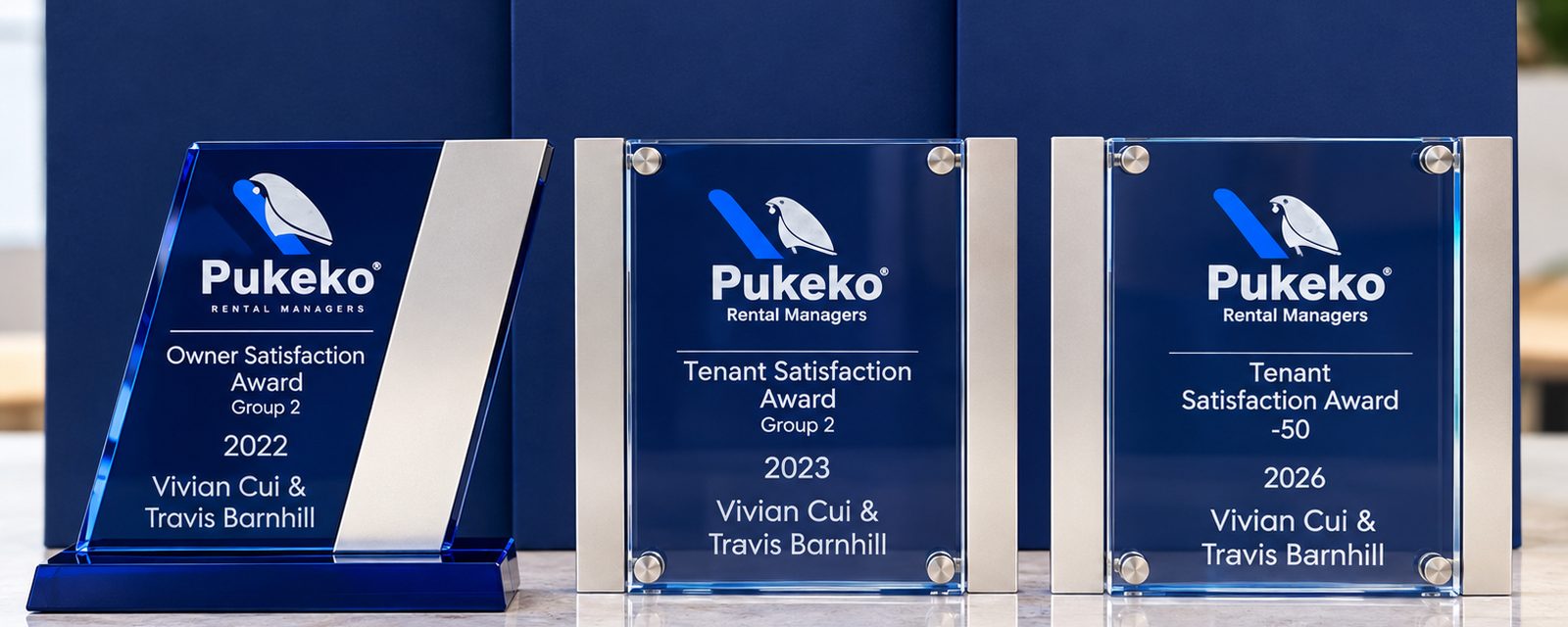 Three blue glass Pukeko Rental Managers awards presented to Vivian Cui and Travis Barnhill, lined up on a marble bench.