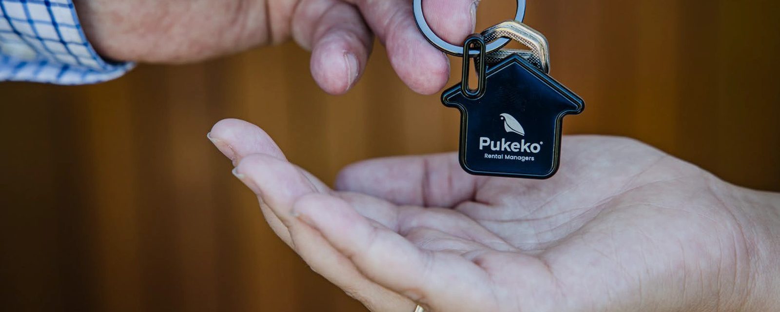 A Pukeko Rental Managers house-shaped keyring being handed over into an open palm, seen close up.