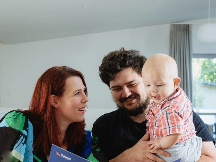 A young couple with a baby reading a blue Pukeko rental management folder at a dining table.
