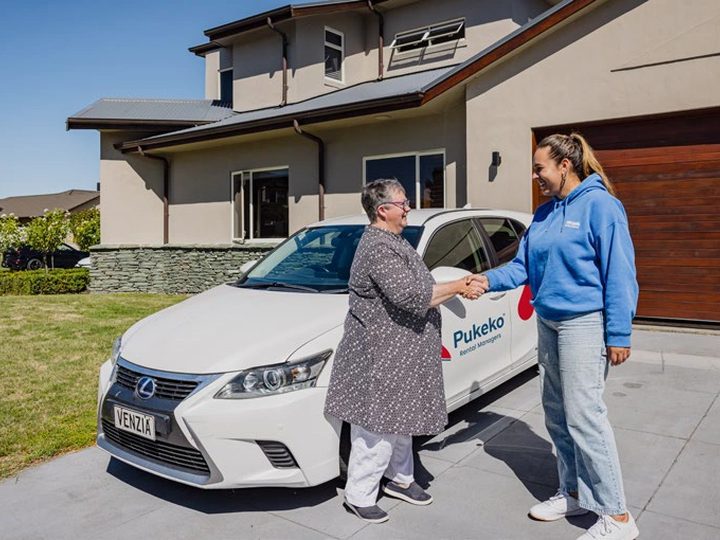 A property owner shaking hands with a Pukeko rental manager beside a Pukeko-branded car in a driveway.