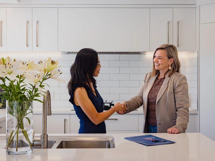 A property owner and a Pukeko rental manager shaking hands across a kitchen bench with a management folder between them.