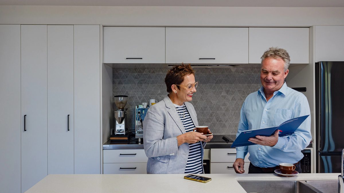 Two Pukeko rental managers talking over coffee in a white kitchen, one holding a blue management folder.